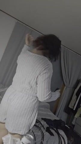 A person is in a dimly lit room and is drying their hair with a hairdryer. The person is wearing a light gray sweater and shorts. There is a bed behind the person. To the right, there is a closet with clothes. In the foreground, there is a can of soda and what appear to be cables.