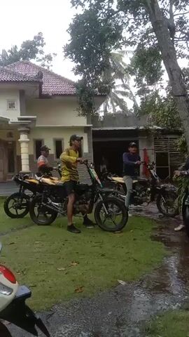 The video shows a group of men gathering on a grassy area in front of a house. Several motorcycles are parked around the group. A man in a blue hat and face covering is loading a large bundle of leafy branches onto his motorcycle. Another man stands with his motorcycle, holding a red bag. The scene appears to be a rural or semi-rural setting with trees and a house in the background.
