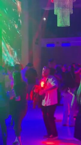 The video shows a crowded indoor space that appears to be a nightclub or party venue. The lighting is dim and colorful, with pink and green lights illuminating the scene. A large screen or projection on the left wall displays abstract, colorful graphics. A multi-tiered chandelier with green lights hangs from the ceiling. People are dancing and mingling in the space. In the foreground, a man in a white shirt and black pants is dancing, holding a black bag over his shoulder. To his left, a woman in a black dress is also dancing. Other individuals are visible in the background, some dancing, some talking, and some standing. The overall atmosphere suggests a lively and energetic event.