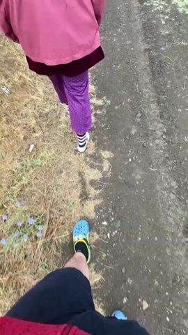 The video begins with a first-person perspective of someone walking on a dirt path, with another person in front wearing a purple jacket and patterned pants. The environment is dry with scattered grass and some small blue flowers. The camera then pans up to show a cloudy sky and a wide, open landscape of dry grass fields, with a dirt road winding through it. Utility poles and power lines are visible in the distance. The video continues to show the same landscape from a slightly different angle, with the person in the purple jacket visible in the distance, walking away from the camera. The weather appears overcast and possibly damp. The video ends with a shot of the landscape, including the power lines.