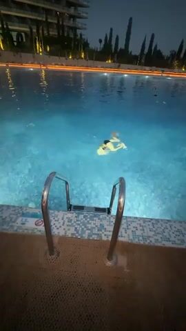 A person is swimming in a brightly lit pool at night. The person is in the middle of the pool, and they appear to be floating on their back. The pool is surrounded by a tiled deck, and there are trees and a building in the background. The building is illuminated with lights, and there are also lights around the edge of the pool. The water in the pool is a clear blue color.