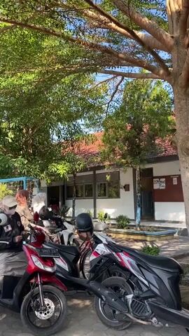 The video shows an outdoor scene in daylight, likely in front of a building that appears to be a school or similar institution. Several motorcycles are parked in the foreground. A red motorcycle is on the left, and a black and white motorcycle is to its right. Two women wearing headscarves are on the black and white motorcycle, with one appearing to operate it. Another person wearing a helmet and a jacket is seated on a red scooter to the far left. The background features a building with a tiled roof and windows, surrounded by trees providing shade. A blue fence is visible to the left of the building. The overall atmosphere seems casual and everyday.