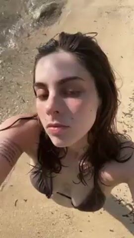 A woman wearing a brown bikini is at the beach. The beach has fine sand. The water is light turquoise with calm waves. She looks up at the camera and raises her arms, adjusting her hair. There is some seaweed on the beach. She turns to the side and looks at the water. In the background are some trees and an island in the distance. The sun is shining brightly.