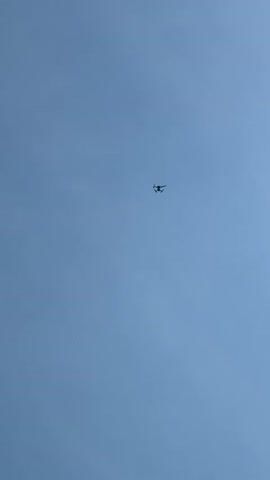 A small, dark quadcopter drone hovers against a clear blue sky. The camera is aimed upwards, capturing the drone as it maintains a fixed position. The sun briefly enters the lower left of the frame, causing a bright lens flare and momentarily silhouetting the drone before moving out of view. The drone remains in the center of the frame against the uniform blue sky for the duration of the video.