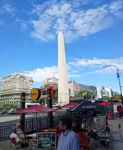 The video shows a cityscape in Buenos Aires, Argentina, with the Obelisco in the background. The sky is bright blue with scattered white clouds. In the foreground, there is a sidewalk with a black wrought-iron fence. Behind the fence, there are outdoor seating areas with red umbrellas and Coca-Cola branding. People are sitting at some of the tables. A man in a blue shirt is standing near the fence. To the left, there is a subway entrance sign with the word "Subte." Across the street, there are several buildings, including one with a large digital billboard and another with ornate architecture. Cars are driving on the street.