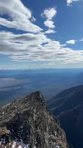 A sweeping panoramic view captures a vast mountain landscape under a bright, partly cloudy sky. The camera pans slowly from left to right, revealing rugged terrain. In the foreground on the left, a sharp, rocky peak with patches of snow juts out prominently, contrasting with a distant, flat valley or plain stretching to the horizon. As the camera continues to pan, a series of dark, forested mountain ridges covered in sparse vegetation and snow patches come into view. Further in the background, a majestic range of snow-capped peaks extends across the horizon under the clear blue sky. The sunlight illuminates the peaks and slopes, casting some areas in shadow. The overall scene depicts a serene and expansive mountainous wilderness, likely in the Arshan region of Russia, characterized by its dramatic elevations and natural beauty. No people or artificial structures are visible.