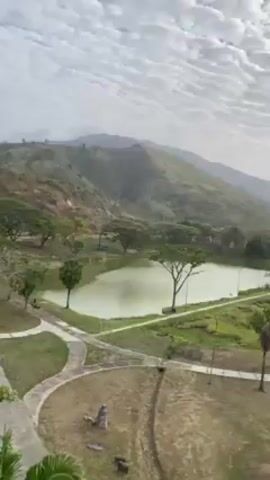 The video shows a serene landscape. In the foreground, there is a man-made lake with a greenish hue, surrounded by trees. A network of paved paths winds through the grassy area surrounding the lake. Beyond the lake, a hillside covered in green vegetation is visible. In the background, there are mountains partially obscured by a slightly overcast sky filled with scattered clouds. There is a person riding a motorcycle in the distance.