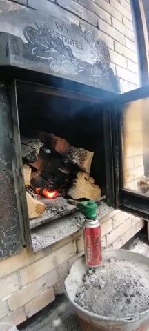 The video shows a fire being started in a wood-burning oven. The oven is made of brick and has a black metal door that is open. Inside the oven, there is a pile of wood logs and burning embers. A portable propane torch with a green nozzle is pointed at the wood to help ignite the fire. A can of "Export Gas" is next to the torch. The floor of the oven is covered with ash and embers. The background is slightly blurred, showing a brick wall and a glimpse of outdoor light. There's smoke visible inside the oven.