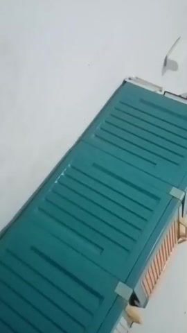 A blurry video shows a teal-colored object with horizontal ridges, possibly a computer component or a toy, on a white surface. The object appears to be a rectangular shape with metal connectors on one end. The background is out of focus and shows a white wall and ceiling with some discoloration. There are glimpses of a round clock and a framed object on the wall. The video quality is poor, with significant blur and low resolution.