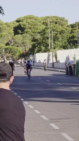 A group of cyclists are participating in a race on a paved road. The cyclists are wearing helmets and athletic attire. Some spectators are visible on the side of the road, observing the race. Trees and buildings line the road, and the sky is clear. The video is shot from a low angle, looking up at the cyclists.