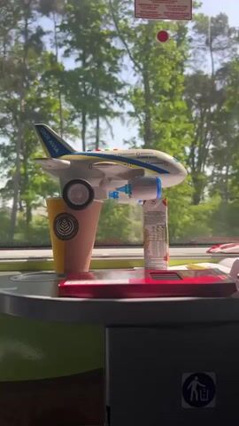 On a moving train, a toy airplane rests precariously on a coffee cup, a juice box acting as a makeshift support. The airplane is branded “UKRAVIA,” suggesting a Ukrainian theme.  The foreground includes a red object, possibly a toy or a game console. Outside the train window, a lush green forest blurs by, indicating the train is traveling through a wooded area. Signage with warning symbols and text is visible at the top of the window.
