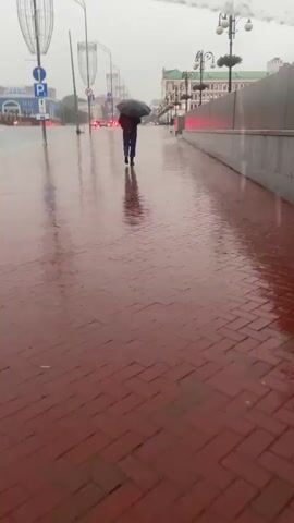 A person walks away from the camera under a black umbrella on a wet, red brick sidewalk during a rainstorm. The sidewalk reflects the overcast sky and city lights. In the background, a street is visible with cars and traffic signals. Buildings line the street, and streetlights are mounted on poles. The rain is visible as streaks falling from above.
