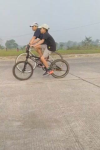 A group of people are riding bicycles and motorcycles on a paved road. The road has white lines and appears to be in a rural or suburban area, with houses and fields visible in the background. The sky is overcast. One person is riding a bicycle and doing a wheelie. Another person is riding a pink and black motorcycle. The video is shaky and seems to be filmed from a moving vehicle.