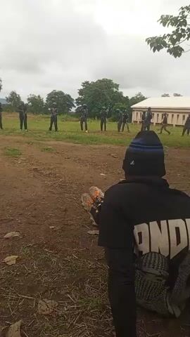 In the foreground, a person wearing a black beanie and a black hoodie with white lettering that appears to say "NDN" is seated on the ground, facing away from the camera. They are wearing black pants with white stripes and grey and white sneakers. In the midground, several individuals in dark uniforms and helmets are standing in a line in a grassy field, holding rifles. They appear to be at a training exercise. In the background, there are trees and a building with a light-colored facade and a white roof. The sky is overcast.