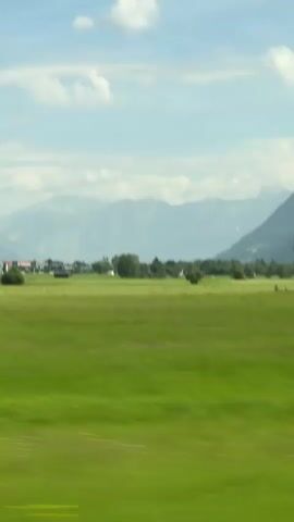 A low-angle, horizontal video captures a blurry, green landscape moving past. In the foreground, the vibrant green grass is heavily blurred, indicating motion. A winding dirt path with tire tracks appears and disappears in the middle ground. Further back, a cluster of buildings, possibly a small village or farm, is visible. Trees dot the landscape between the buildings and the fields. In the distance, hazy, layered mountains form the horizon under a pale blue sky with scattered white clouds. The overall impression is of traveling through a rural, mountainous region.