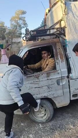 A man in a gray hoodie and black pants, who is wearing a black beanie and gloves, stands on the driver's side of a beat-up white van. He holds his hands out as he talks to the driver, a man wearing glasses and a brown jacket. The van has a rack on top and a white canvas covering the back. The background includes trees and a building, and the ground is covered in dirt and gravel.