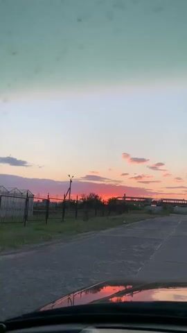 This is a vertical, first-person perspective video from inside a vehicle driving on an asphalt road. The road curves slightly to the right. On the left side of the road, there is a grassy area with a fence and some trees. Beyond the fence, there are some low-lying structures, possibly under construction, with a crane visible to the right of them. The sky is a gradient of colors, with a pale green at the top transitioning to pink and orange near the horizon, indicating a sunset. The setting sun is visible as a bright orange glow behind the structures. The hood of the vehicle is visible at the bottom of the frame, reflecting the colors of the sunset. The video shows movement as the vehicle progresses along the road, with the landscape and sky changing slightly.