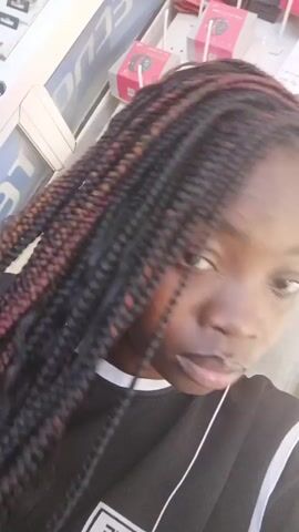 A young Black woman is centered in the frame, taking a selfie. She has dark eyes and braided hair, a few of which have red accents. She is wearing a black t-shirt with a white design at the bottom. A white earbud wire is visible. She briefly runs her right hand through her hair. Behind her is a shelf stocked with various items, including a sign with the word "ECHO" visible.