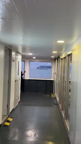 This video shows the interior of a ferry or boat. A person wearing black clothing and white sneakers is standing and looking out a window to the left. In the center, there is a large rectangular window with a view of the sea and a distant boat. Another person wearing a dark jacket and black pants is leaning against the right side of the large window. The interior has a dark floor, white ceiling with circular lights, and white walls with metallic accents and doors. The overall lighting suggests it might be dusk or dawn.