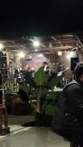 A band performs on a stage at night. The band consists of at least four members: a male singer with glasses and a white shirt, a female singer, a male guitarist in a suit, and another male guitarist. They are illuminated by stage lights. In the foreground, there is a large green plant and patrons are seated at tables, with one person in a black puffer jacket facing away from the camera. The stage is covered by a rustic, open-air roof structure with visible lighting fixtures. The background appears to be a wall with some signage. The overall atmosphere seems to be a casual outdoor dining or entertainment venue.