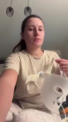 Here is a description of the video:

A woman sits on a bed, wearing patterned pajamas and a light-colored t-shirt. She is putting on a white plastic face mask. The mask has cutouts for the eyes, nose, and mouth. It also has straps on the sides. A white wire extends from the mask. In the background, there is a baby chair, a wooden structure, and two pendant lights.