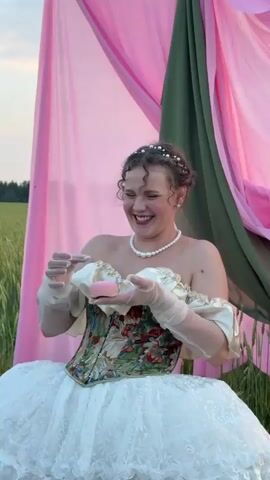 A woman wearing a white ball gown and a corset is in a field. She is laughing and holding a small pink object, possibly a bar of soap, in her gloved hands. Pink and green fabric drapes behind her. The scene appears to be outdoors during the day, with a clear sky visible.