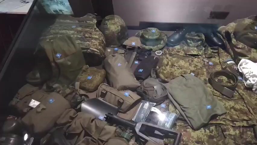 The video shows a display of military equipment and mannequins dressed in camouflage uniforms. The first scene is a top-down view of various pieces of military gear laid out, including helmets, vests, bags, clothing, boots, and a shovel. The gear is arranged neatly in a display case. The subsequent scenes show mannequins in military uniforms positioned in a diorama-like setting, with backdrops depicting natural environments like forests and rocky terrain. One mannequin is shown in a crouching position, aiming a rifle, while another is in a similar pose. The mannequins are dressed in full camouflage attire, including helmets, body armor, and tactical vests. The overall impression is that of a museum exhibit showcasing military equipment and uniforms.