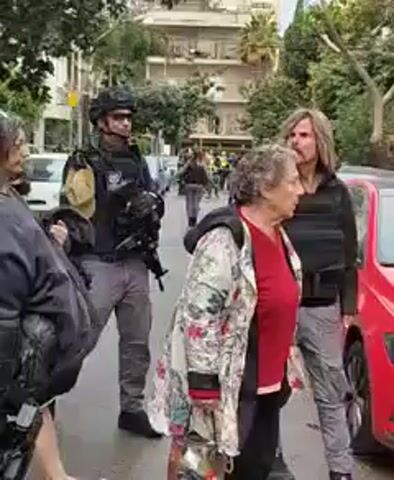 The video shows an outdoor street scene. Multiple people are present, including several uniformed law enforcement officers, equipped with tactical gear and carrying weapons. One officer is wearing a helmet and goggles. Some civilians are also visible, including women and a man with long hair. Cars are parked along the street. The setting appears to be an urban environment with buildings and trees in the background. The overall scene suggests a potentially tense situation.