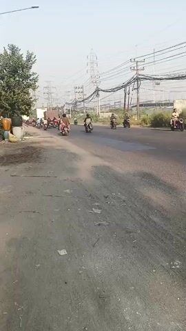 A street scene with several motorcycles visible. The road is paved and dusty, with a dirt shoulder. Vegetation and some structures are on the side of the road, and power lines are overhead. One motorcycle is in the foreground with a rider, while others are further down the road. The weather appears to be clear and sunny.