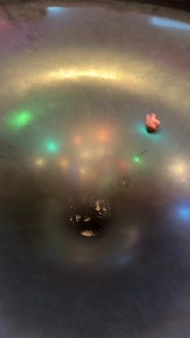 The video shows a close-up, overhead view of a dark, circular surface, possibly a metal bowl or basin, with several blurred colored lights reflecting on its surface. At the center of the surface, there is a dark circular depression, and within this depression, a small, bright green object is visible. To the left of this depression, a small object with red and white stripes, appearing to be a spinning top or a marble, is in motion, creating a blur of color. The objects are spinning rapidly, and the movement suggests a dynamic process occurring within the depression. The lighting conditions are dim, with the colored reflections being the primary source of illumination besides the central green object.