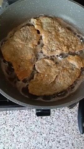 The video shows three pieces of breaded meat being fried in a pan on a stovetop. The meat is golden brown and appears to be crisping in hot oil. Bubbles are visible around the meat, indicating that it is actively cooking. The pan is gray and non-stick, and it is sitting on a speckled countertop. A black stovetop burner is partially visible beneath the pan.