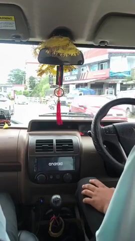 The video shows the interior of a car from the perspective of the passenger. The dashboard is visible, featuring an Android infotainment system, air vents, and a gear shift. A hand is resting on the steering wheel. Outside the car, through the windshield, a street scene is visible with other vehicles, buildings, and trees. The car appears to be in motion.