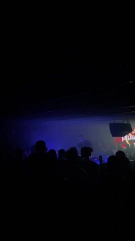 The video shows a dimly lit club with people silhouetted in the foreground, facing a DJ on a stage. The stage has a screen displaying graphics. Lighting shifts from yellow to blue throughout the video. Attendees hold up phones, seemingly recording the performance. The venue has a low ceiling with visible white beams.