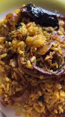A plate of jollof rice with fish is shown in a close-up shot. The rice is yellowish in color and has some green vegetables mixed in. The fish is arranged on top of the rice and looks dark brown. The plate itself is yellow and blue. The lighting appears to be from above. The focus is on the food. There is some faint music playing in the background.