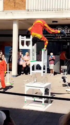 A male performer in a red and yellow costume is upside down, balancing on his right hand on top of a stack of three white chairs. His legs are extended upwards. The stack of chairs is placed on a white table, which is also on the ground. There are several glass bottles on the table beneath the chairs. The performer is outdoors, in front of a brick building with a white balcony. Several people are watching the performance, including a woman in a pink shirt and a man in a light blue shirt. A camera on a tripod is visible to the right of the performer.