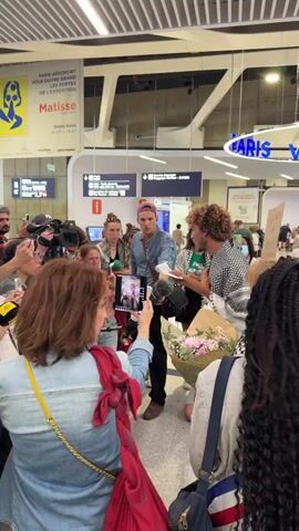 The scene unfolds inside an airport terminal, likely in Paris, as indicated by signage. A small crowd has gathered, comprised of individuals of various ages and ethnic backgrounds, some holding cameras, microphones, and mobile phones, seemingly focused on two men. One of the men, with curly hair, is wearing a patterned black-and-white jacket. The other man wears a denim shirt and a baseball cap. He is holding something that appears to be a white stick or scroll. The men are surrounded by people eager to capture the moment. The crowd includes women with various hairstyles, one with a red scarf, and others with braided hair. There is a bouquet of flowers. Airport signs display directions and information about a Matisse exhibition. The overall atmosphere suggests a public appearance or arrival of the two men.