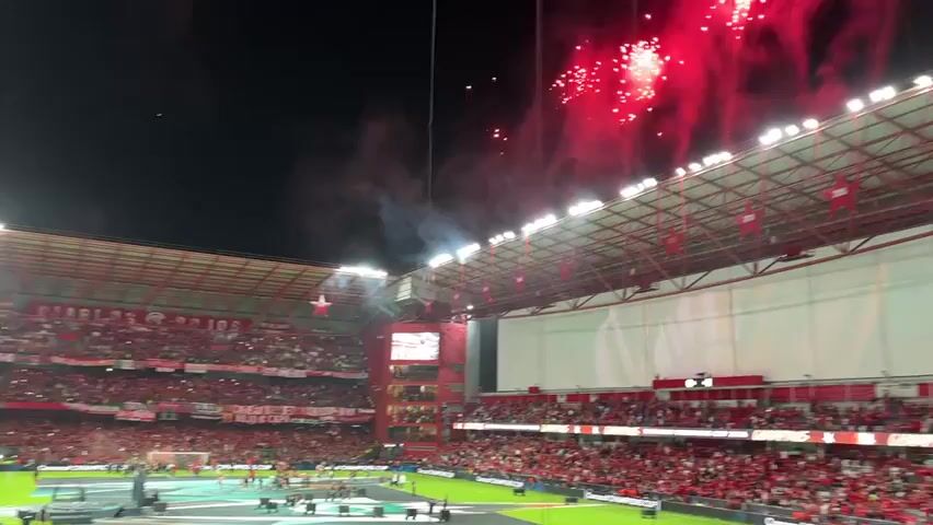 The video shows a nighttime view of a packed soccer stadium with fireworks going off overhead. The stands are filled with people, many wearing red, and banners can be seen hanging from the upper levels. The stadium is well-lit, and a large screen is visible. The field is green, and players are present. The atmosphere appears celebratory, with the crowd cheering and the fireworks adding to the excitement.