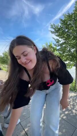 A young woman with long brown hair, wearing a black off-the-shoulder top with a pink flower accessory and light-wash jeans, is bending over and laughing while looking at her phone, which she holds in her left hand. She is outdoors, near a body of water, with trees and a blue sky with clouds in the background. Another person, wearing grey pants and a white shirt, is partially visible on the left side of the frame, sitting down. The ground is paved with light-colored stones.