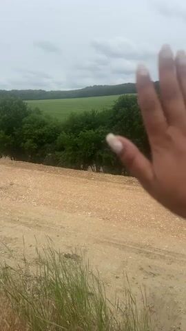The video shows a landscape featuring a dirt road in the foreground, with green vegetation and trees in the mid-ground, and a field or meadow stretching to the horizon under a cloudy sky. A person's hand is partially obscuring the view. The foreground shows dry, gravelly soil and some tall grass. The mid-ground consists of lush green trees and a flat expanse that may be a field or cultivated area. The background showcases a distant tree line against a cloudy sky.