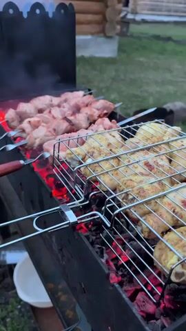 The video captures an outdoor grilling scene, likely during late afternoon or early evening. In the foreground, a black metal brazier holds glowing red coals, emitting wisps of smoke. Several metal skewers are laden with pieces of raw or lightly cooked meat, possibly pork or lamb (shashlik), positioned directly over the hot coals. Adjacent to the skewers, a metal grill basket contains what appears to be chicken drumsticks or pieces, also cooking over the embers. In the background, green grass extends towards a rustic wooden log house or cabin, suggesting a rural or countryside setting consistent with Tyulyuk, Russia. No people are visible, but the focus is entirely on the cooking process.