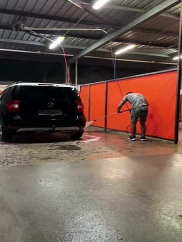 A person in a grey t-shirt and grey shorts is using a hose to spray down the rear of a black SUV parked at a self-service car wash. The car wash has an open-air roof with overhead fluorescent lights. Orange tarps divide the car wash bays. The ground is wet and reflective. The license plate of the car is partially visible.
