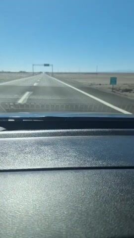 The video shows a view from the inside of a car, looking out through the windshield at an empty highway stretching towards the horizon under a clear blue sky. The dashboard of the car is visible in the foreground, and the highway is marked with white lines. A large overhead sign is visible in the distance. The landscape on either side of the highway appears to be dry and barren, suggesting a desert or arid region. There are no other vehicles or people visible in the frame. The sun is bright, casting a glare on the dashboard. The video appears to be shot while the car is in motion, as indicated by the slight blur of the passing landscape.