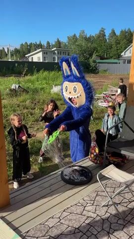 A person in a blue furry costume is blowing bubbles outdoors with children. The costume appears to be a cartoon character with a large head and blue fur. The person is holding a bubble wand, and multiple bubbles are floating in the air. Several children are present, some of whom are reaching out to touch the bubbles. The scene takes place on a wooden deck with a view of a grassy yard and houses in the background. The sky is clear and blue, indicating a sunny day.
