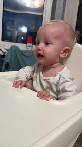 A baby with blond hair sits in a white highchair. The baby is wearing a white, long-sleeved shirt with a bear print. The baby looks off to the left and opens its mouth to make sounds. A dark window is behind the baby with a dark blue curtain. A red box is sitting on a table near the window.