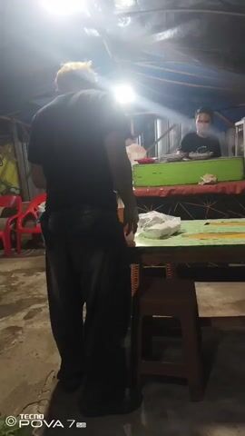 The video shows a dimly lit indoor setting, possibly a small eatery or shop. In the foreground, a person wearing a black shirt and dark pants stands with their back to the camera. They have light-colored hair. A table with a green surface is positioned in front of them. In the background, a person wearing a dark shirt and a mask is visible behind a counter. The counter has a green section and appears to be part of a food stall. There are lights illuminating the area. The environment seems rustic, with exposed ceiling structures and possibly tarpaulin overhead. The footage is shaky and slightly blurry.