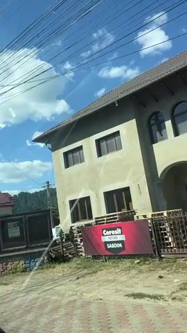 The video shows a view from inside a vehicle traveling along a street. On the left side of the street, there is a tall utility pole with numerous wires and two bicycles attached. Further down the pole, there is a green trash can. Adjacent to the pole is a building with a stone and brick facade and three dark-framed display cases on top. Beyond that, there are trees and more buildings. On the right side of the street, there are houses with pitched roofs, fences, and trees. The sky is blue with scattered white clouds, and sunlight is visible. The street has a paved surface and a sidewalk with a patterned brick design. Reflections on the vehicle's windshield are also visible.