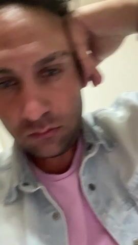 A man is filmed from the chest up as he rests his head on his hand, looking into the camera. He is wearing a light blue denim jacket over a pink t-shirt. The background appears to be a room with wallpaper. The camera briefly pans and tilts, creating a shaky effect before the video ends.