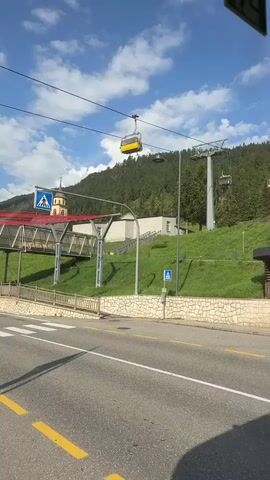 A yellow cable car moves across the frame, suspended by wires against a backdrop of a lush green, tree-covered mountain under a bright blue sky with white clouds. In the foreground, a paved road runs horizontally, marked with yellow lines. To the left, a pedestrian bridge with a red roof spans over the road. Behind the bridge, a building with a tall, slender tower and a pointed roof, resembling a church, stands out. Several smaller buildings are visible further up the grassy slope. A modern ski lift tower and more cables are present on the right side of the frame, also leading up the mountain. A bus stop shelter with benches is situated near the road on the right. The scene appears to be a resort or mountain town during the daytime.