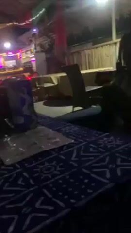 The video shows an indoor scene with a table in the foreground, covered with a blue patterned cloth. A metal cup, a menu, and a blue box are on the table. In the background, there's a bar area with illuminated shelves filled with bottles and other bar equipment. There are people in the background, but they are not clearly visible. The lighting is dim, with various colored lights illuminating the bar area and the ceiling decorations. The ceiling appears to have a thatched design. The camera appears to be stationary.