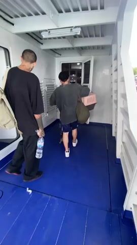 The video shows two men walking out of an enclosed area on a boat onto an open deck. The man in front is wearing a black t-shirt and black pants, carrying a beige bag over his shoulder and a water bottle in his hand. The other man is wearing a dark t-shirt and shorts. The boat is docked at a pier. The video then pans to show the surrounding ocean, other boats, and a green island in the distance under a sky with scattered clouds. The video ends on a shot of the ocean from the back of the boat.